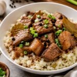 Pork Adobo served over cauliflower rice in a white bowl with garlic, bay leaves, and green onions.